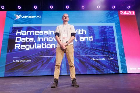 Image of keynote speaker Phil Winder with the title slide of the presentation behind him.