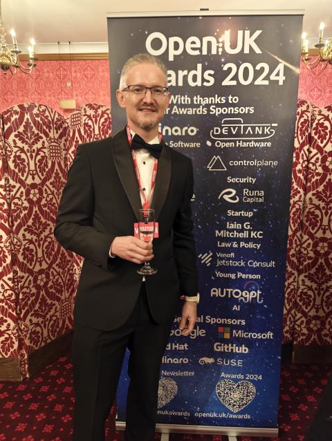 Phil Winder in black tie beside the OpenUK Awards 2024 sponsor banner in the Cholmondeley Room